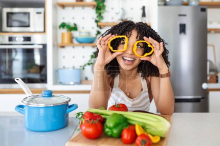Foto de Foto de mujer joven sonriendo mientras cocina ensalada con verduras frescas en el interior de la cocina en casa - Imagen libre de derechos