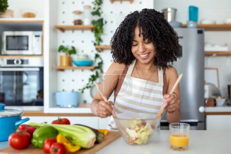 Foto de Feliz sonrisa linda mujer está preparando una ensalada vegana fresca y saludable con muchas verduras en la cocina en casa y probar una nueva receta - Imagen libre de derechos