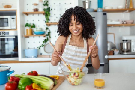 Téléchargez les photos : Jeune femme souriante mignonne abondante sur la cuisine prépare une salade végétalienne en vêtements décontractés. - en image libre de droit