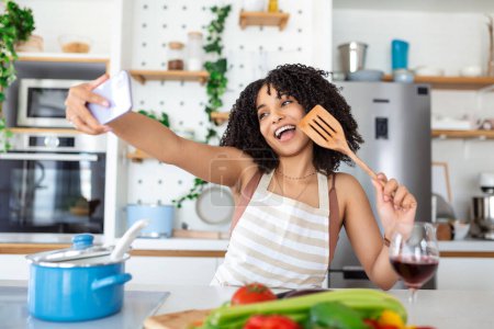 Foto de Retrato de una joven afroamericana sonriente tomando selfie con smartphone mientras cocina en casa - Imagen libre de derechos