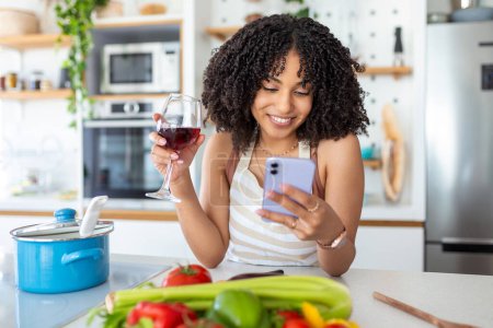 Foto de Mujer sosteniendo el teléfono móvil y una copa de vino en la cocina en casa - Imagen libre de derechos