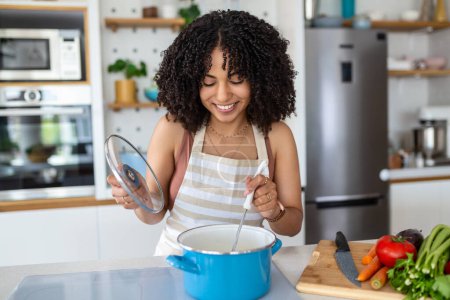 Foto de Happy African American Woman Cooking Cena de degustación en una olla de pie en la cocina moderna en casa. Ama de casa preparando comida sana sonriendo a la cámara. Hogar y Nutrición. Concepto de recetas de dieta - Imagen libre de derechos