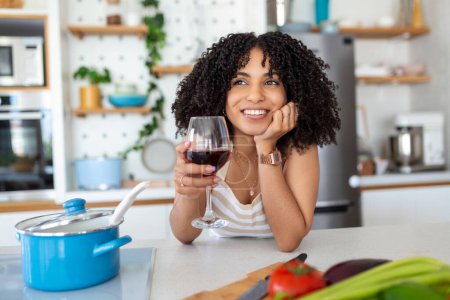 Foto de Sonriente joven ama de casa con copa de vino tinto mirando a la cámara mientras ella está parada en la estufa en la cocina preparando la cena - Imagen libre de derechos
