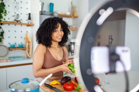 Foto de Feliz mujer afroamericana sonriendo y demostrando verduras maduras mientras filma video para cocinar vlog en la cocina en casa - Imagen libre de derechos