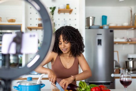 Photo for Professional beautiful happy young woman is blogging for her kitchen channel about healthy living in the kitchen of her home and looking on camera on a tripod - Royalty Free Image