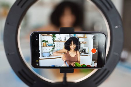Foto de Mujer afroamericana chef vlogger disparar cocina comida vlog blog en el teléfono inteligente en la cocina casera. Mujer joven grabar receta o tutorial en el teléfono celular preparando plato vegetariano saludable. - Imagen libre de derechos
