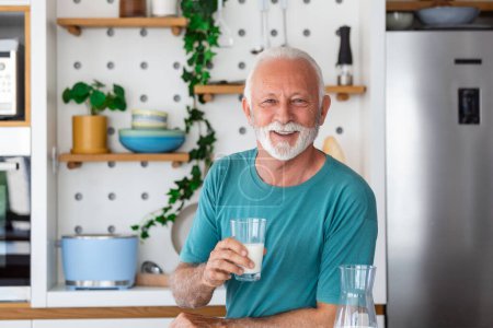 Foto de Hombre mayor bebiendo un vaso de leche con una cara feliz de pie y sonriendo. Hombre mayor guapo bebiendo un vaso de leche fresca en la cocina - Imagen libre de derechos