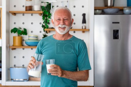 Foto de Hombre mayor bebiendo un vaso de leche con una cara feliz de pie y sonriendo. Hombre mayor guapo bebiendo un vaso de leche fresca en la cocina - Imagen libre de derechos