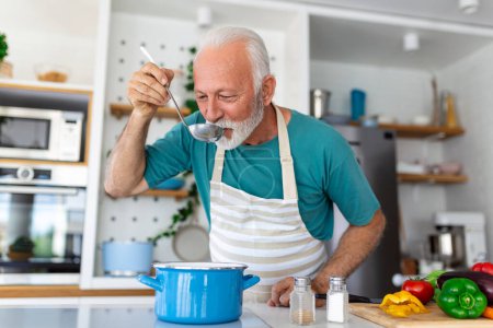 Téléchargez les photos : Joyeux retraité cuisinier dans la cuisine. Retraite, concept de hobby people. Portrait d'un homme âgé souriant tenant une cuillère pour goûter à la nourriture - en image libre de droit