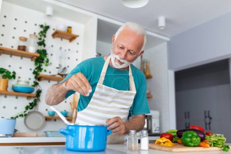 Foto de Feliz hombre mayor divirtiéndose cocinando en casa - Anciano preparando almuerzo de salud en cocina moderna - Tiempo de vida jubilado y concepto de nutrición alimentaria - Imagen libre de derechos