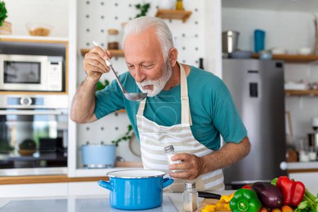 Téléchargez les photos : Joyeux retraité cuisinier dans la cuisine. Retraite, concept de hobby people. Portrait d'un homme âgé souriant tenant une cuillère pour goûter à la nourriture - en image libre de droit