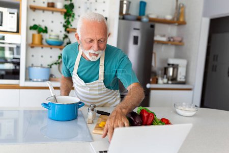 Foto de Feliz hombre mayor divirtiéndose cocinando en casa - persona mayor preparando un almuerzo saludable en la cocina moderna mirando el recibo en su computadora portátil - Imagen libre de derechos