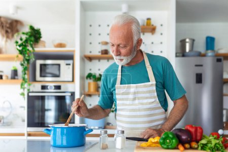 Foto de Feliz hombre mayor divirtiéndose cocinando en casa - Anciano preparando almuerzo de salud en cocina moderna - Tiempo de vida jubilado y concepto de nutrición alimentaria - Imagen libre de derechos