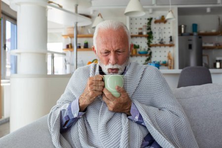 Foto de Hombre mayor que sufre de gripe tomando té mientras está sentado envuelto en una manta en el sofá en casa. Hombre mayor enfermo con dolor de cabeza sentado debajo de la manta en la sala de estar. Hombre con un resfriado acostado en el sofá sosteniendo una taza de té caliente. - Imagen libre de derechos