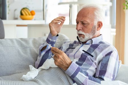 Photo for Sick elderly man checking his temperature suffering from seasonal flu or cold lying on sofa caughing suffering from seasonal flu or cold. . Ill senior feel unhealthy with influenza at home - Royalty Free Image
