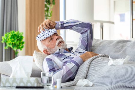 Foto de Hombre maduro sintiéndose enfermo de resfriado y fiebre en casa, enfermo de gripe sentado en el sofá con bolsa de hielo en la cabeza - Imagen libre de derechos