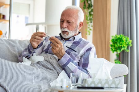 Sick elderly man checking his temperature suffering from seasonal flu or cold lying on sofa caughing suffering from seasonal flu or cold. . Ill senior feel unhealthy with influenza at home