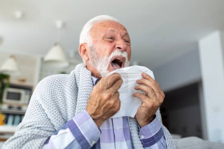 Photo for Senior sick bearded man sneezes into napkin at home on gray sofa with white blanket. Disease, protection, coronavirus, virus, disease, flu, respiratory dressing. - Royalty Free Image