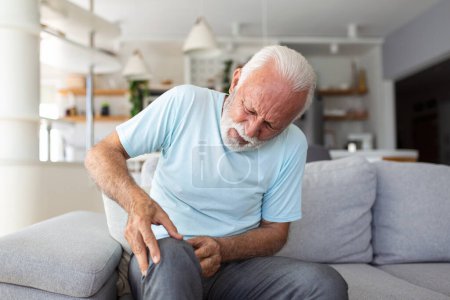 Foto de El hombre mayor está sentado en el sofá en casa, tiene dolor en la articulación de la rodilla, sosteniendo su pierna, concepto de osteoartritis. - Imagen libre de derechos