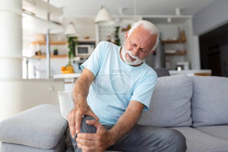 Téléchargez les photos : L'homme plus âgé est assis sur le canapé à la maison, a des douleurs dans l'articulation du genou, tenant sa jambe, concept d'arthrose. - en image libre de droit