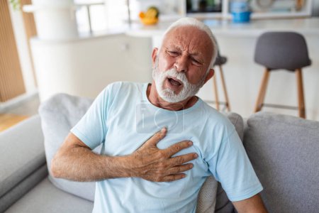 Photo for Senior man presses hand to chest has heart attack suffers from unbearable pain, Mature man with pain on heart in living room. Senior man suffering from bad pain in his chest heart attack at home - Royalty Free Image