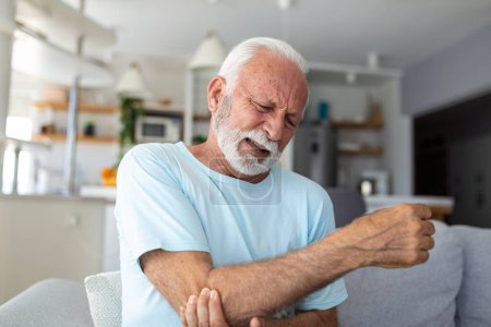 Senior man with arm pain.Old male massaging painful hand indoors. Old man hand holding his elbow suffering from elbow pain. Senior man suffering from pain in hand at home. Old age, health