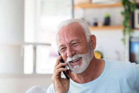 Téléchargez les photos : Homme âgé parlant sur un téléphone portable à la maison. Rire homme âgé parlant sur un téléphone portable tout en se relaxant dans une chaise dans son salon - en image libre de droit