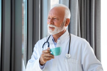 Photo for Portrait of senior mature health care professional, doctor, with stethoscope holding blue cup of coffee, - Royalty Free Image