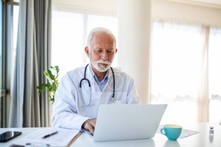 Photo for Serious mature doctor using laptop and sitting at desk. Senior professional medic physician wearing white coat and stethoscope working on computer at workplace. - Royalty Free Image