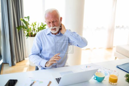 Foto de El escote. Hombre mayor tocando el cuello que sufre de dolor sentado trabajando en el ordenador portátil en la oficina moderna. Atención médica, problemas de salud en la vejez Concepto - Imagen libre de derechos