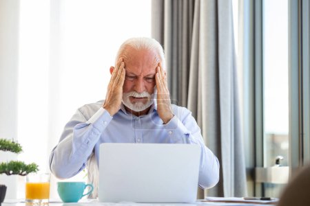 Téléchargez les photos : Homme d'affaires mature stressé avec ordinateur portable. Il pourrait aussi avoir mal à la tête. Il est assis dans la salle de réunion. Il y a des documents et un ordinateur portable sur la table. Espace de copie - en image libre de droit
