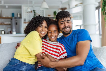 Photo for Love And Care. Portrait of cheerful family of three people hugging sitting on the sofa at home. Smiling young girl embracing her parents. - Royalty Free Image