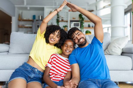 Foto de Concepto de Cuidado Familiar, Protección y Seguro. Retrato de padres afroamericanos sonrientes haciendo techo simbólico de manos por encima de su hija feliz, sentados en la alfombra del suelo en la sala de estar en casa - Imagen libre de derechos