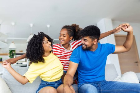 Cheerful african mother and father playing with daughter at home. Cute girl enjoying sitting on father shoulder while looking at camera. Young fmily having fun together on the sofa at home.