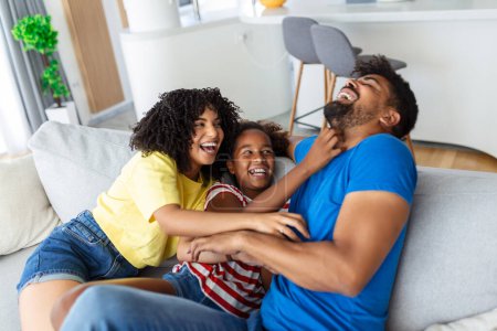 Fröhliche Menschen, die auf der Couch im Wohnzimmer sitzen, haben Spaß kleine Tochter kitzelnde Mutter lacht zusammen mit Eltern genießen Freizeit zu Hause spielen. Wochenendaktivitäten glücklich Familie Lifestyle-Konzept