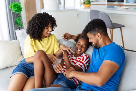 Foto de Feliz familia multiétnica sentados en el sofá riendo juntos. Padres alegres jugando con su hija en casa. Padre hace cosquillas a su niña mientras la madre sonríe. - Imagen libre de derechos