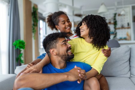Foto de Amor y cuidado. Retrato de familia alegre de tres personas abrazándose sentadas en el sofá en casa. Sonriente jovencita abrazando a sus padres. - Imagen libre de derechos