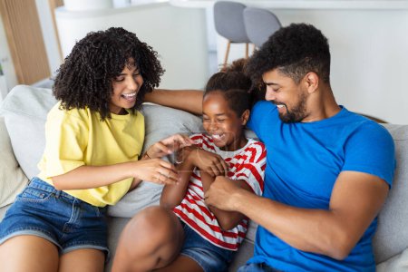 Photo for Happy multiethnic family sitting on sofa laughing together. Cheerful parents playing with their daughter at home. Father tickles his little girl while the mother is smiling. - Royalty Free Image