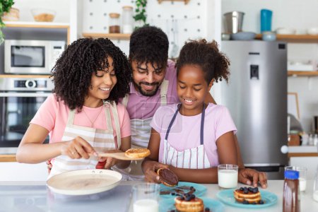 Photo for Glad positive young family foolishes with small daughter at kitchen, making pancakes, have tasty breakfast, pose at camera, drink fresh milk, eat pancakes - Royalty Free Image
