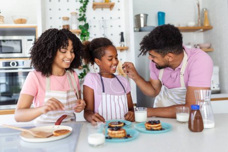 Foto de Preparando panadería casera. Amar a los cónyuges familiares afro-americanos amigables con su hija cocinando alimentos juntos en la mesa de la cocina, haciendo panqueques caseros - Imagen libre de derechos
