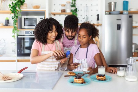 Téléchargez les photos : Famille à la maison Manger le petit déjeuner dans la cuisine ensemble, Happy African American famille debout autour de la cuisinière, fille manger des crêpes - en image libre de droit