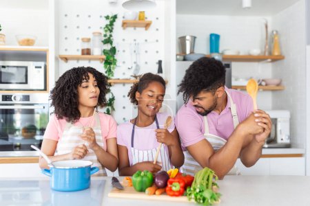 Téléchargez les photos : Joyeux jeunes parents et petite fille s'amusent tout en cuisinant ensemble dans la cuisine, heureux afro-américains famille chantant et dupant, en utilisant la spatule et fouetter comme microphones, copier l'espace - en image libre de droit