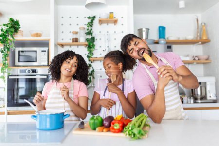 Foto de Familia afroamericana juguetona divirtiéndose en la cocina, cantando y bailando mientras cocina el almuerzo en casa, familia alegre con Daugther usando espátulas como micrófonos, espacio libre - Imagen libre de derechos