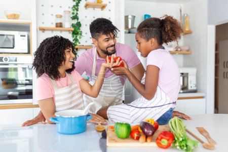 Téléchargez les photos : Petite fille mignonne et ses beaux parents sourient tout en cuisinant dans la cuisine à la maison. Heureuse famille afro-américaine préparant ensemble des aliments sains dans la cuisine - en image libre de droit