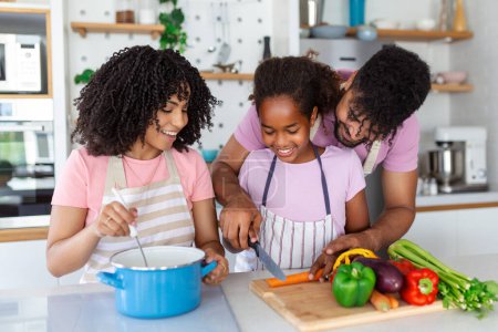 Téléchargez les photos : Petite fille mignonne et ses beaux parents coupent des légumes et sourient tout en cuisinant dans la cuisine à la maison - en image libre de droit