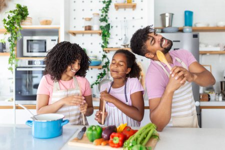 Téléchargez les photos : Cuisine Fun. Portrait de parents afro-américains positifs et petite fille chantant et dansant tout en cuisinant ensemble à la cuisine, utilisant la spatule comme microphone, préparant le repas - en image libre de droit