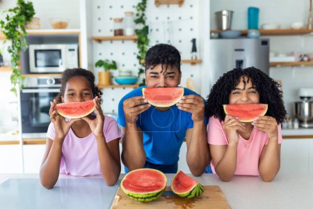 Photo for Portrait of cheerful young family taking a bite of a watermelon. African American family standing together at a kitchen eating watermelon. - Royalty Free Image
