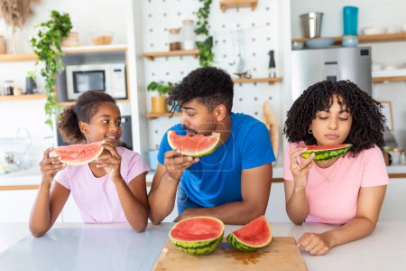 Téléchargez les photos : Portrait d'une jeune famille joyeuse prenant une bouchée de pastèque. Famille afro-américaine debout ensemble dans une cuisine mangeant pastèque. - en image libre de droit