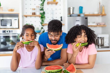 Photo for Young Family eating watermelon and having fun. Mixed race family in kitchen together eating a watermelon slice. - Royalty Free Image
