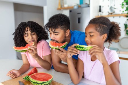 Photo for Young Family eating watermelon and having fun. Mixed race family in kitchen together eating a watermelon slice. - Royalty Free Image